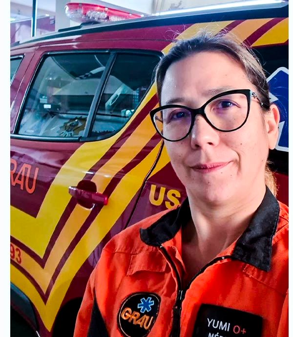 Dra. Yumi em ambiente de atendimento de urgência, em frente a uma ambulância